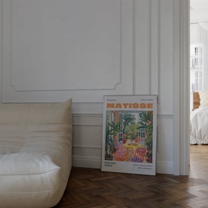 May include: A white couch with a white throw pillow sits in front of a white wall with a framed poster of a painting by Henri Matisse. The poster features a colorful painting of a garden scene with a table and chairs. The poster is titled "Matisse" and includes the text "06200 Nice, France" and "Explore Matisse's World #TheMatisseCollection".