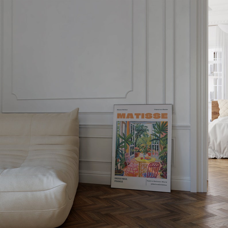 May include: A white couch with a white throw pillow sits in front of a white wall with a framed poster of a painting by Henri Matisse. The poster features a colorful painting of a garden scene with a table and chairs. The poster is titled "Matisse" and includes the text "06200 Nice, France" and "Explore Matisse's World #TheMatisseCollection".