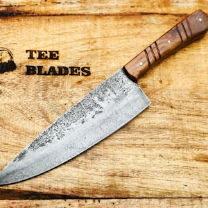 May include: A handmade chef's knife with a rustic, textured blade and a wooden handle. The knife is displayed on a wooden cutting board with the text "TEE BLADES" and an eagle logo.