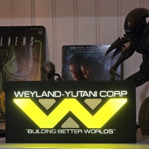 May include: A display of collectibles from the Alien franchise. Includes a glowing Weyland-Yutani Corp sign, a Xenomorph figure, a power loader model, and movie packaging.