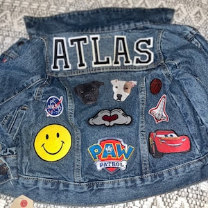 Puede incluir: Una chaqueta vaquera azul con el nombre "ATLAS" bordado en la espalda. La chaqueta está decorada con varios parches, incluyendo una cara sonriente, un logotipo de la patrulla canina, un cohete, un coche y un perro.