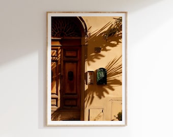 May include: A wooden door with a decorative archway and a metal door knocker. The door is partially shaded by a palm tree, casting a shadow on the yellow wall. There are two mailboxes on the wall.