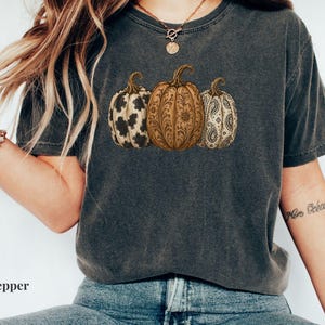 May include: A charcoal grey short-sleeve t-shirt featuring a graphic print of three pumpkins. The left pumpkin has a black and white cow print, the center pumpkin has a brown floral pattern, and the right pumpkin has a black and white paisley design.