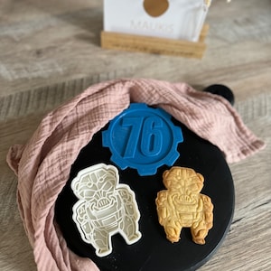 Könnte beinhalten: Ein blauer Plastik-Ausstecher in Form eines Zahnrads mit der Zahl 76 darin. Es gibt zwei weiße Plastik-Ausstecher in Form von Bären. Ein Bären-Ausstecher liegt neben einem braunen Bären-Keks.