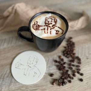 Könnte beinhalten: Eine schwarze Kaffeetasse mit einem weißen Schaumdesign einer Person, die einen Daumen nach oben zeigt. Die Tasse steht auf einer Holzoberfläche mit einem weißen Untersetzer und verstreuten Kaffeebohnen.