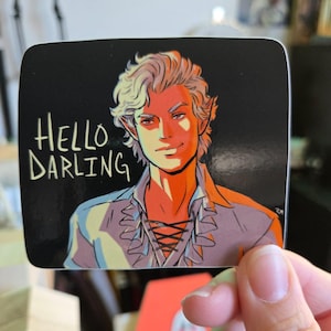Puede incluir: Una pegatina con una ilustración de dibujos animados de una persona con cabello rubio y una camisa de color morado claro. La persona sonríe y el texto "Hello Darling" está escrito en blanco sobre un fondo negro.