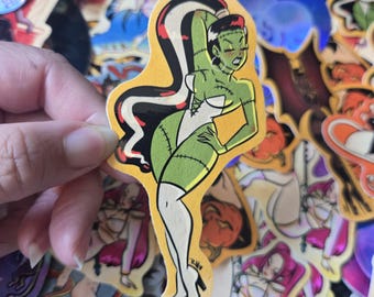 May include: A sticker depicting a cartoon monster woman with green skin, black and white hair, and a white outfit. The sticker has a yellow background and is surrounded by other stickers.