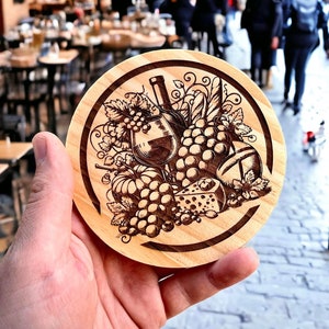 May include: A wooden coaster with a laser-engraved design of grapes, cheese, bread, and a glass of wine. The design is in shades of brown and black.