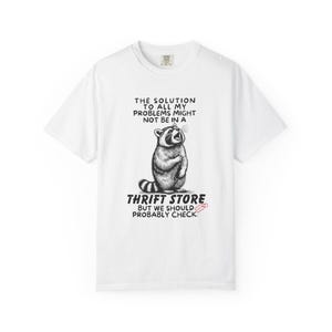 Flat lay photo of soft relaxed fit white shirt featuring a mischievous raccoon illustration with the text, The solution to all my problems might not be in a thrift store, but we should probably check. Giving off playful, feral raccoon meme vibes. Perfect for gifting to your thrifting partner in crime or rocking yourself on your next bargain hunt. Available on white, ivory or grey.