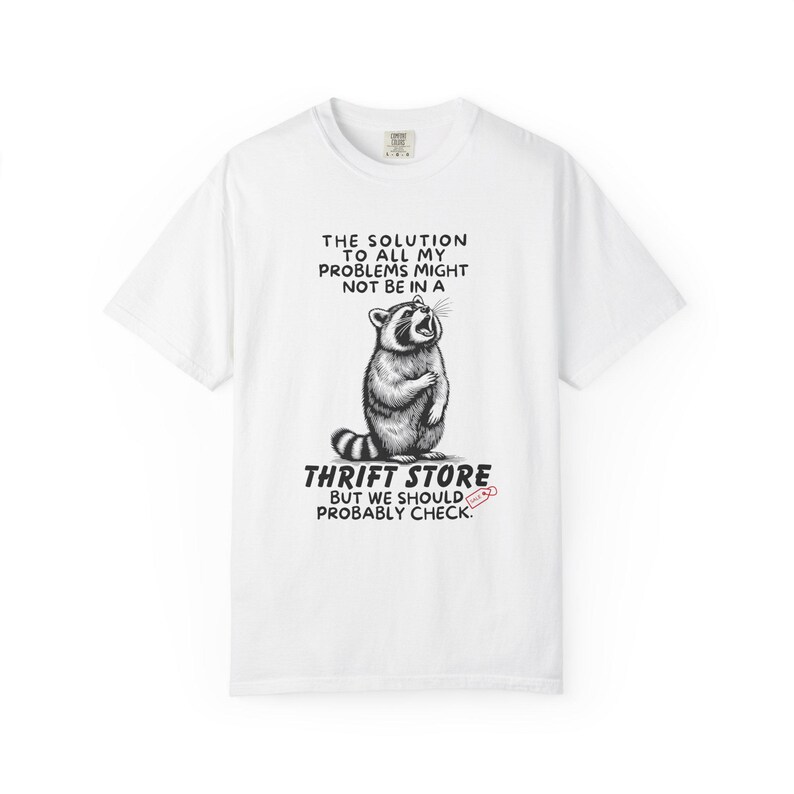 Flat lay photo of soft relaxed fit white shirt featuring a mischievous raccoon illustration with the text, The solution to all my problems might not be in a thrift store, but we should probably check. Giving off playful, feral raccoon meme vibes. Perfect for gifting to your thrifting partner in crime or rocking yourself on your next bargain hunt. Available on white, ivory or grey.