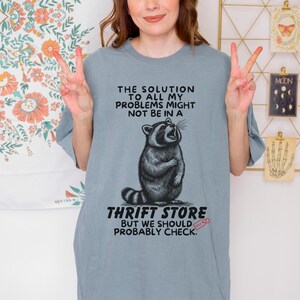 Photo of model wearing soft relaxed fit grey shirt featuring a mischievous raccoon illustration with the text, The solution to all my problems might not be in a thrift store, but we should probably check. Giving off playful, feral raccoon meme vibes. Perfect for gifting to your thrifting partner in crime or rocking yourself on your next bargain hunt. Available on white, ivory or grey.