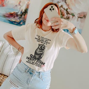 Photo of model wearing soft relaxed fit ivory shirt featuring a mischievous raccoon illustration with the text, The solution to all my problems might not be in a thrift store, but we should probably check. Giving off playful, feral raccoon meme vibes. Perfect for gifting to your thrifting partner in crime or rocking yourself on your next bargain hunt. Available on white, ivory or grey.