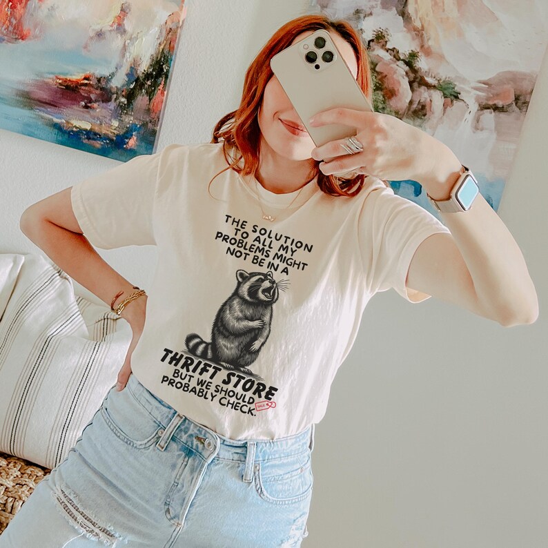 Photo of model wearing soft relaxed fit ivory shirt featuring a mischievous raccoon illustration with the text, The solution to all my problems might not be in a thrift store, but we should probably check. Giving off playful, feral raccoon meme vibes. Perfect for gifting to your thrifting partner in crime or rocking yourself on your next bargain hunt. Available on white, ivory or grey.