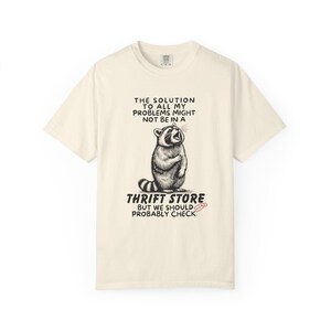 Flat lay photo of soft relaxed fit ivory shirt featuring a mischievous raccoon illustration with the text, The solution to all my problems might not be in a thrift store, but we should probably check. Giving off playful, feral raccoon meme vibes. Perfect for gifting to your thrifting partner in crime or rocking yourself on your next bargain hunt. Available on white, ivory or grey.