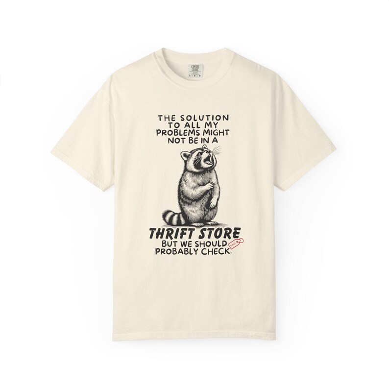 Flat lay photo of soft relaxed fit ivory shirt featuring a mischievous raccoon illustration with the text, The solution to all my problems might not be in a thrift store, but we should probably check. Giving off playful, feral raccoon meme vibes. Perfect for gifting to your thrifting partner in crime or rocking yourself on your next bargain hunt. Available on white, ivory or grey.