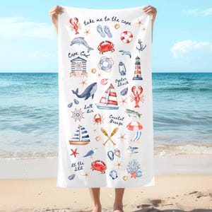 Puede incluir: Toalla de playa blanca con ilustraciones náuticas, incluyendo un faro, un velero, una langosta y estrellas de mar. El texto incluye "take me to the cape", "Cape Cod", "Salt Air" y "Coastal Breeze". La toalla se sostiene contra un fondo de playa y océano.