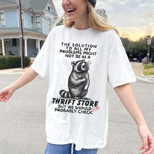 Photo of model wearing soft relaxed fit white shirt featuring a mischievous raccoon illustration with the text, The solution to all my problems might not be in a thrift store, but we should probably check. Giving off playful, feral raccoon meme vibes. Perfect for gifting to your thrifting partner in crime or rocking yourself on your next bargain hunt. Available on white, ivory or grey.