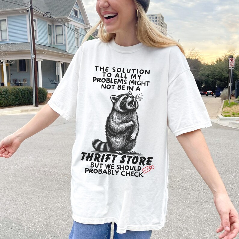 Photo of model wearing soft relaxed fit white shirt featuring a mischievous raccoon illustration with the text, The solution to all my problems might not be in a thrift store, but we should probably check. Giving off playful, feral raccoon meme vibes. Perfect for gifting to your thrifting partner in crime or rocking yourself on your next bargain hunt. Available on white, ivory or grey.