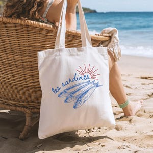May include: A white canvas tote bag with a blue and red design featuring three sardines and the text "les sardines". The bag is sitting on a sandy beach.