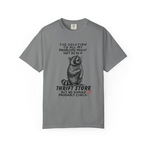 Flat lay photo of soft relaxed fit grey shirt featuring a mischievous raccoon illustration with the text, The solution to all my problems might not be in a thrift store, but we should probably check. Giving off playful, feral raccoon meme vibes. Perfect for gifting to your thrifting partner in crime or rocking yourself on your next bargain hunt. Available on white, ivory or grey.