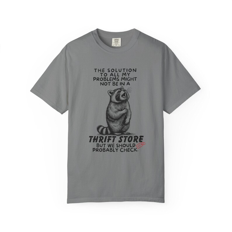 Flat lay photo of soft relaxed fit grey shirt featuring a mischievous raccoon illustration with the text, The solution to all my problems might not be in a thrift store, but we should probably check. Giving off playful, feral raccoon meme vibes. Perfect for gifting to your thrifting partner in crime or rocking yourself on your next bargain hunt. Available on white, ivory or grey.