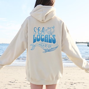 Könnte beinhalten: Ein cremefarbener Hoodie mit einem blauen Aufdruck auf der Rückseite. Der Aufdruck zeigt einen Wal mit Blumen, eine Meeresschildkröte und den Text "SEA the LOCALS".