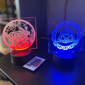 May include: Two illuminated acrylic night lights with a remote control. The left light features a red glow and a white silhouette of a rabbit in a circle. The right light features a blue glow and a white Celtic knot design in a circle.