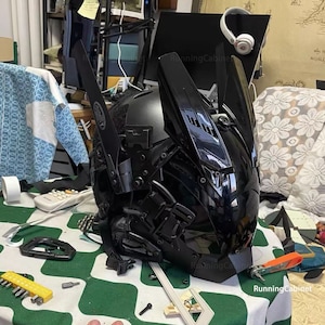 May include: A black helmet with a futuristic design and multiple moving parts. The helmet has a black visor and is made of a hard plastic material.