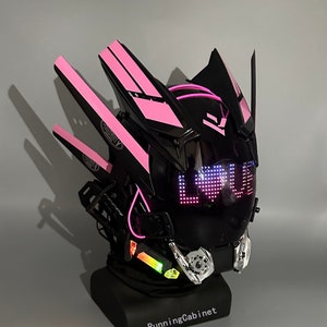 Op de afbeelding: Een zwarte en roze motorhelm met een vizier dat het woord "LOVE" in roze LED-verlichting weergeeft. De helm heeft een futuristisch ontwerp met scherpe hoeken en een verscheidenheid aan details.