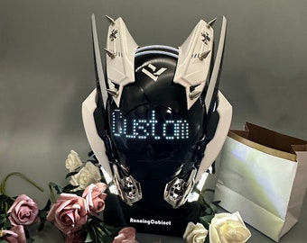 May include: A white and black helmet with a custom LED display that reads "Custom" on the front. The helmet has two large white wings with spikes on the top. The helmet is sitting on a black stand with the words "RunningCabinet" printed on it. There are pink and white roses in the background.