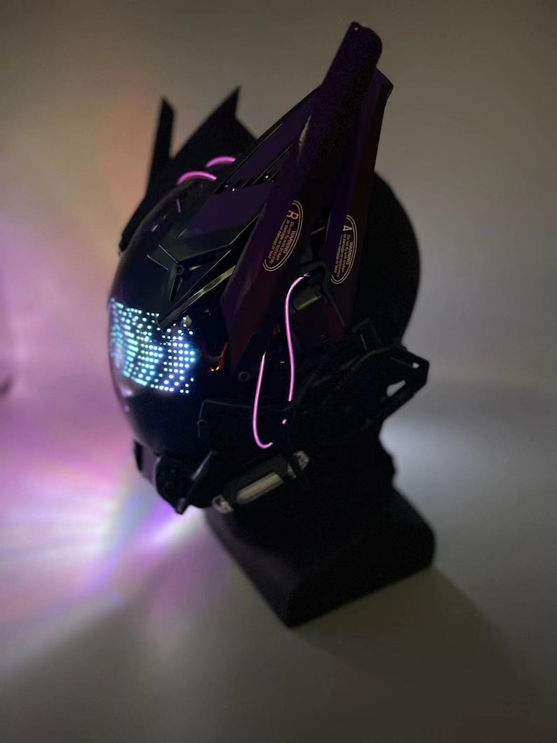 Op de afbeelding: Een zwarte helm met een paars en zwart ontwerp en een verlicht display aan de voorkant. De helm staat op een zwarte standaard.