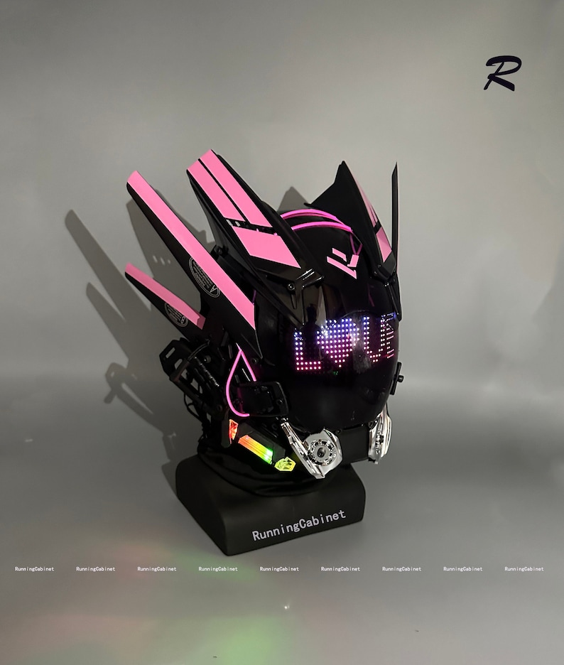 Op de afbeelding: Een zwarte en roze motorhelm met een vizier dat het woord "LOVE" in roze LED-verlichting weergeeft. De helm heeft een futuristisch ontwerp met scherpe hoeken en een verscheidenheid aan details.