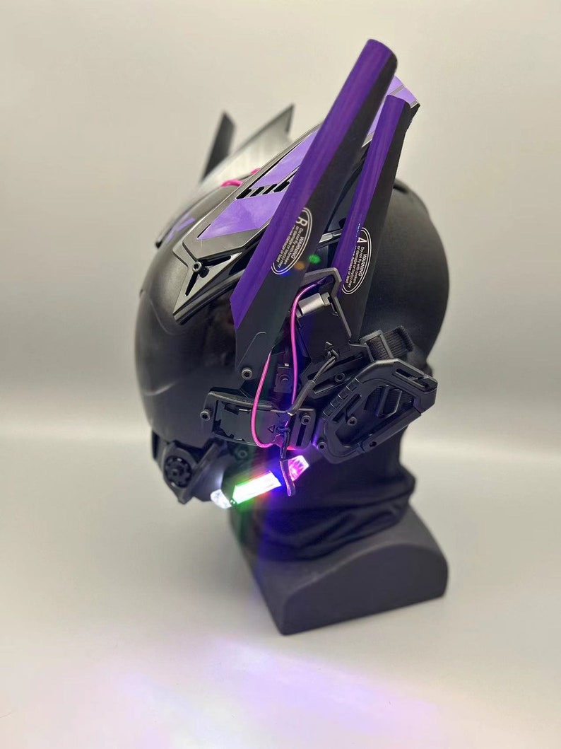 Op de afbeelding: Een zwarte helm met paarse accenten en een verlichtingsfunctie. De helm heeft een futuristisch ontwerp en staat op een zwarte standaard.