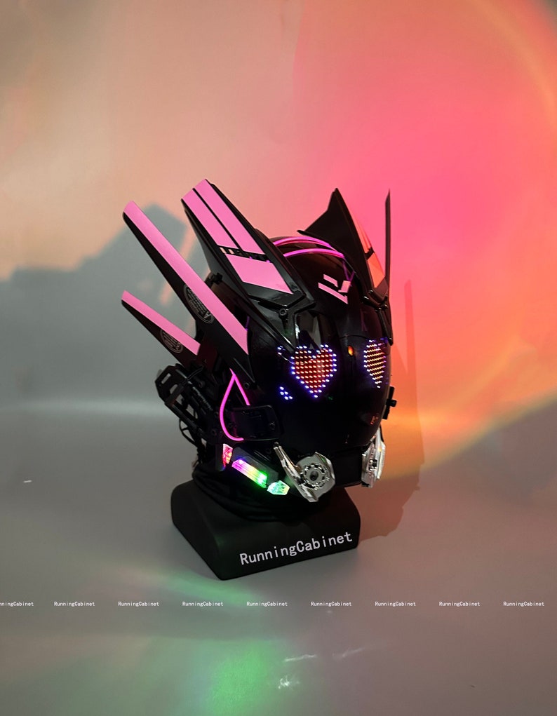 Op de afbeelding: Een zwarte en roze helm met een hartvormig LED-display. De helm heeft een futuristisch ontwerp met scherpe hoeken en een vizier. Het LED-display licht op in de vorm van een roze hart.