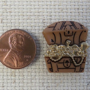 May include: A brown plastic treasure chest charm filled with gold coins and jewelry. A US penny is next to the charm for size comparison. The chest has black detailing and a lock. The charm is on a beige textured surface.