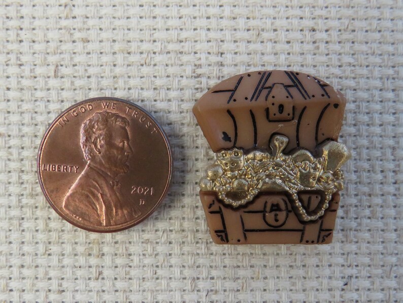 May include: A brown plastic treasure chest charm filled with gold coins and jewelry. A US penny is next to the charm for size comparison. The chest has black detailing and a lock. The charm is on a beige textured surface.