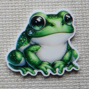May include: A sticker featuring a cartoon frog with large, expressive eyes. The frog is primarily green and white, with detailed shading and highlights. The sticker has a white border and is set against a neutral background.