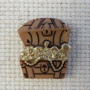 May include: A tan treasure chest brooch with a hinged lid, filled with gold coins and jewellery. The chest has black outline details and a keyhole. The treasure inside is gold-coloured.