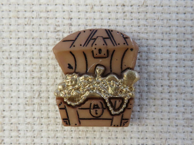 May include: A tan treasure chest brooch with a hinged lid, filled with gold coins and jewellery. The chest has black outline details and a keyhole. The treasure inside is gold-coloured.