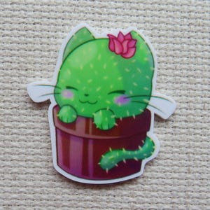 May include: A whimsical sticker featuring a green cactus cat in a brown pot. The cat has a pink flower on its head, pink cheeks, and a smiling face. The sticker is on a neutral background.