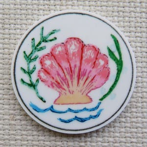 May include: A round, white decorative item featuring a pink seashell design. The seashell is surrounded by green seaweed and blue waves. The item has a black border.