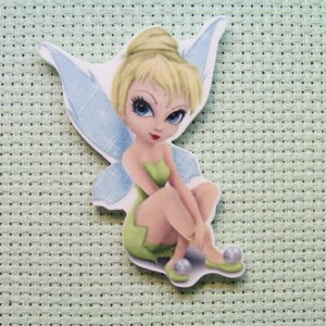 May include: A Tinkerbell fairy figurine with blue wings, blonde hair, and a green dress. She is sitting with her legs crossed and has a sweet expression on her face.