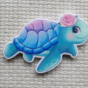 May include: A whimsical sticker of a cartoon turtle with a blue and purple shell and a light blue body. The turtle has a pink rose on its head and rosy cheeks. The sticker is on a textured, off-white surface.