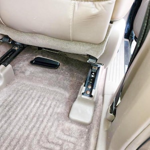 May include: A beige leather vehicle seat with a storage pocket on the back, positioned above a brown carpeted floor. The seat is mounted on a black metal track mechanism with visible circular adjustment holes and a plastic cover at the base.
