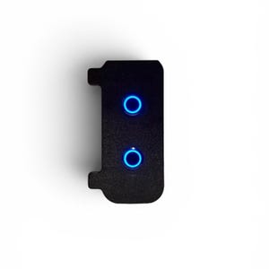 May include: A rectangular black control panel with a textured surface featuring two circular push buttons. Each button is illuminated by a bright blue ring light. The panel has small protruding tabs on the left side for mounting purposes.