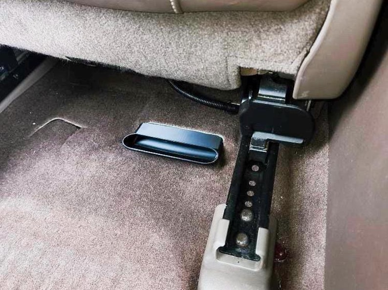 Puede incluir: Un compartimento de almacenamiento de pl&aacute;stico negro est&aacute; unido a la parte posterior de un asiento de coche. El compartimento est&aacute; abierto y parece estar vac&iacute;o.
