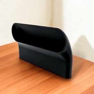 Puede incluir: Un organizador de pl&aacute;stico negro con forma curva y un solo compartimento. El organizador est&aacute; dise&ntilde;ado para guardar objetos como bol&iacute;grafos, l&aacute;pices u otros objetos peque&ntilde;os.
