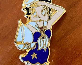 Op de afbeelding: Een gouden speld met een cartoonfiguur in een matrozenpak. Het personage is een vrouw met zwart haar, een blauwe jurk, een witte matrozenpet en witte kousen. Ze houdt een klein wit zeilbootje vast.