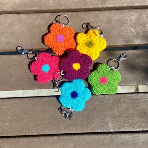 May include: Six colorful flower-shaped keychains with a silver ring. The keychains are made of yarn and have a 3D puffy texture. The colors are orange, yellow, pink, purple, green, and blue.