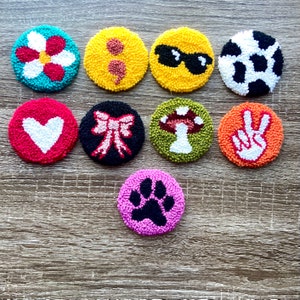 May include: Ten colorful round felt circles with different designs, including a flower, sunglasses, a peace sign, a paw print, a heart, a mushroom, a bow, and a cow print.